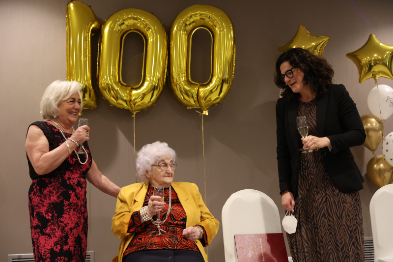Starsza kobieta elegancko ubrana siedzi na fotelu, obok kobieta w eleganckiej sukni z mikrofonem i młodsza kobieta, w tle napis sto lat