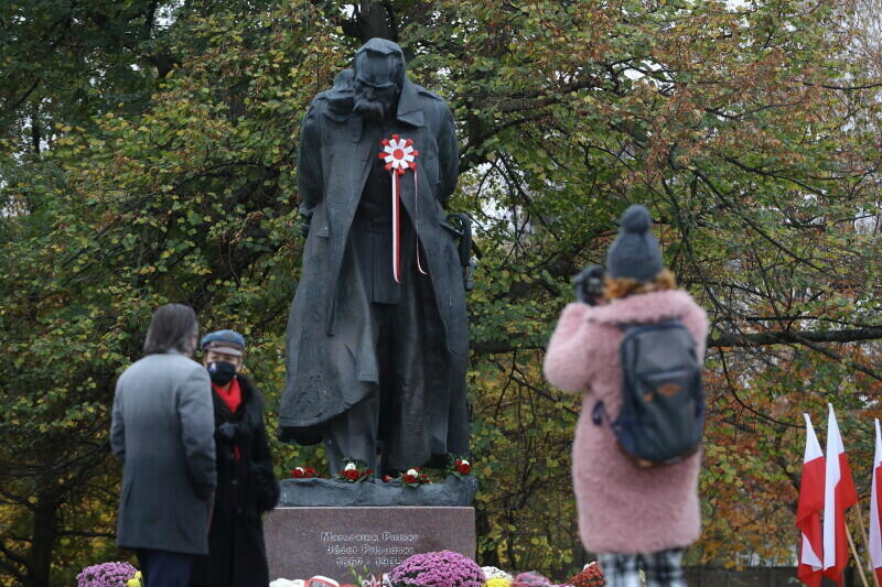 Zdjęcie przedstawia widok pomnika Piłsudskiego od przodu. Przed pomnikiem stoją trzy osoby, jedna z nich robi zdjęcie. Pomnik na piersi postaci marszałka ozdobiony jest dużym biało-czerwonym kotylionem. Przed pomnikiem stoją kwiaty, po prawej widać biało-czerwone flagi 