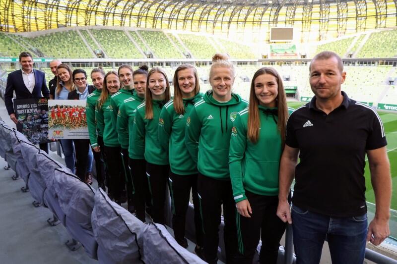 Stadion Gdańsk Arena, na trybunach stoją młode dziewczęta w strojach sportowych, jedna za drugą. Z przodu mężczyzna w średnim wieku, z tyłu kobieta w średnim wieku z długimi włosami 