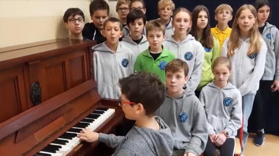 Grupa uczniów stoi przy pianienie, na pianinie gra uczeń 