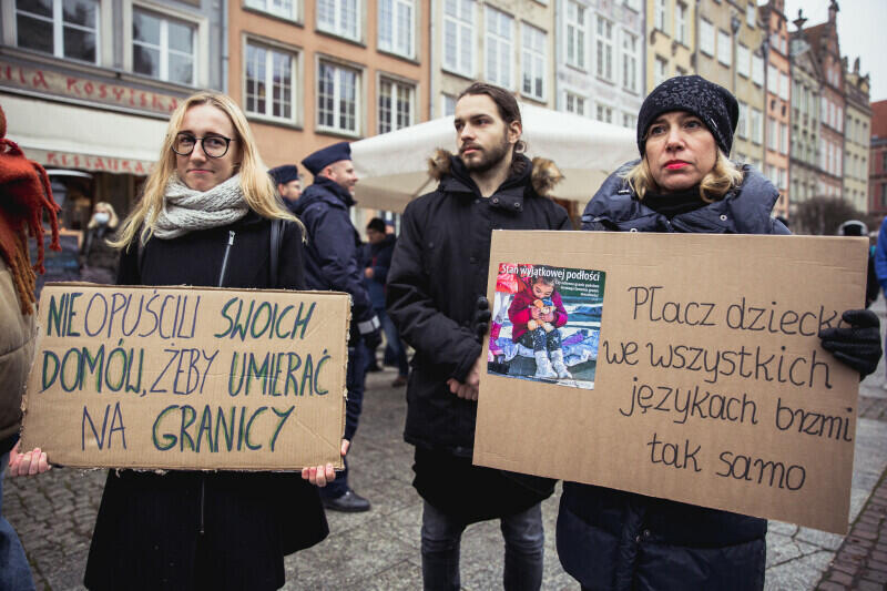 Zdjęcie z manifestacji na Długim Targu w sobotę, 13 listopada 2021 roku. Na pierwszym planie stoją trzy osoby: kobieta, mężczyzna i kobieta. Kobiety trzymają hasła wypisane na dwóch transparentach z kartonu: "Nie opuścili swoich domów, żeby umierać na granicy" i "Płacz dziecka we wszystkich językach brzmi tak samo"