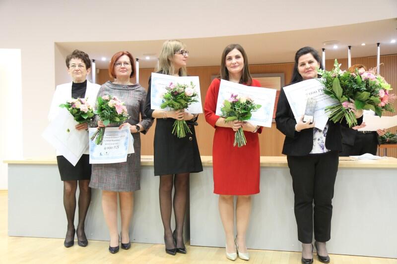 Cztery kobiety w różnym wieku, elegancko ubrane, stoją z kwiatami i dyplomami w ręku 