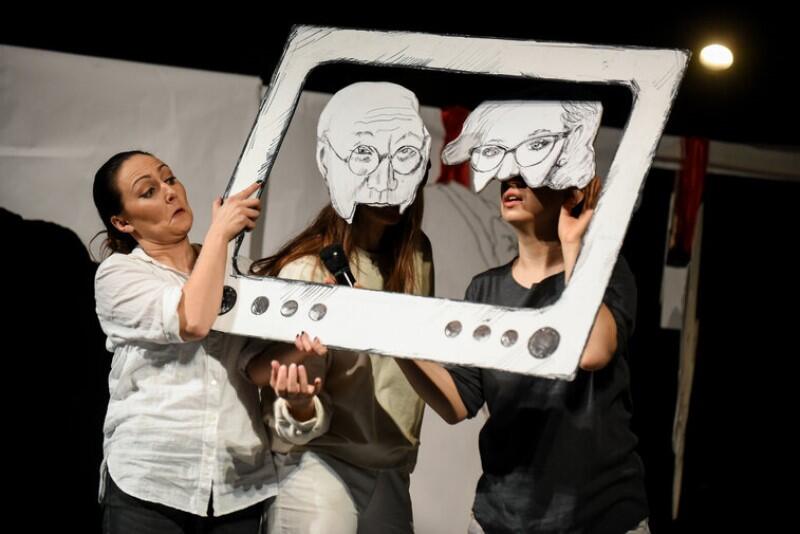 Scena teatralna, na niej stoi kobieta w średnim wieku, trzyma dekorację przedstawiającą ekran telewizora. W środku telewizora sa dwie postacie kobieta i mężczyna 