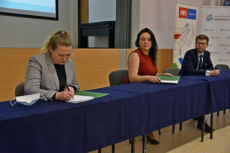 dwie kobiety i jeden mężczyzna w garniturze siedzą za stołem, jedna z kobiet podpisuje dokument