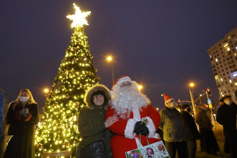Rozświetlona białymi światełkami choinka, przed nią stojący święty Mikołaj i grupa osób