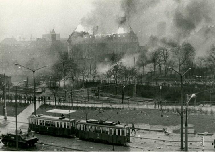 czarno białe zdjęcie,  tramwaj , na ulicy milicja i mnóstwo porozrzucanych kameni, w oddali płonie budynek