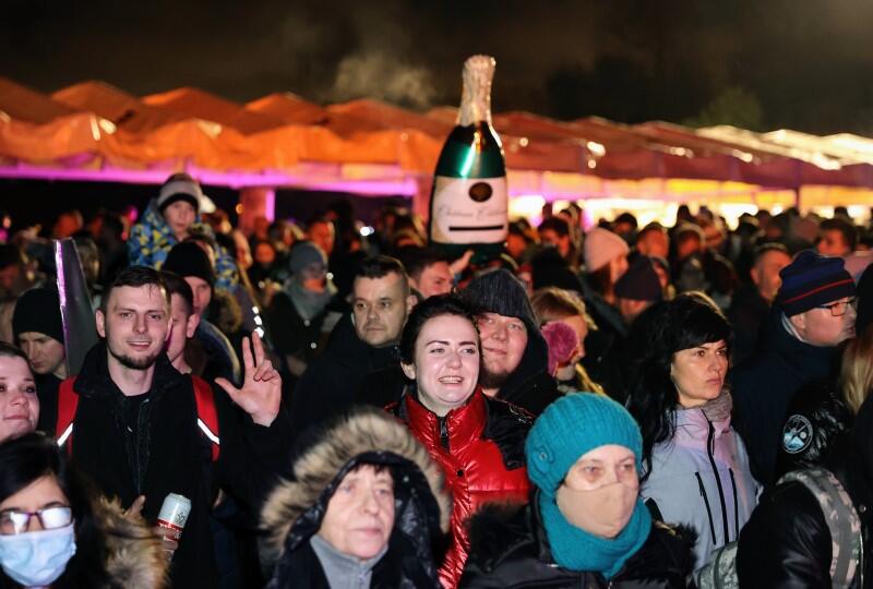 tłum uśmiechniętych ludzi, nad którymi widnieje duża butelka szampana