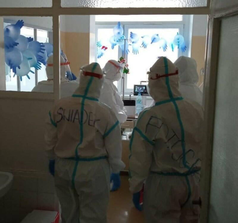 Sala szpitalna, drzwi, tyłem stoją postacie w uniformach sanitarnych 