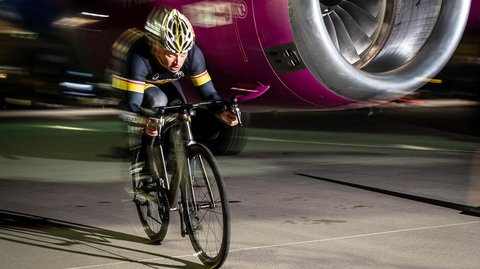 rowerzysta w stroju sportowym pędzi na rowerze nocą, w tle silnik samolotu