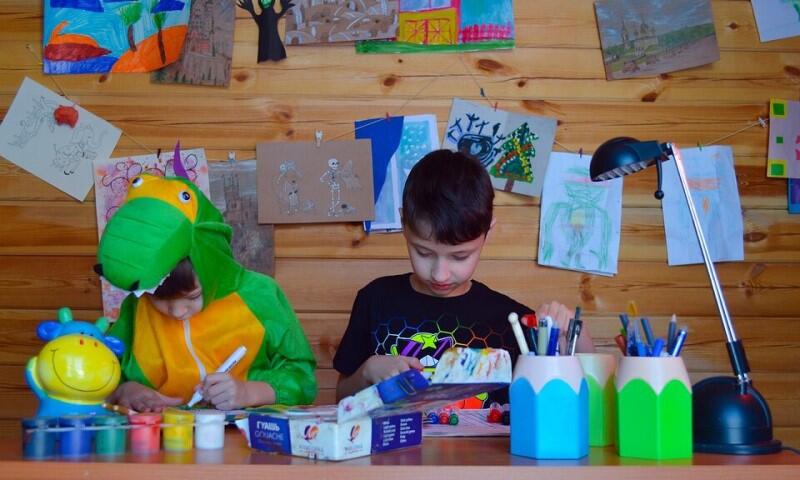 Dwoje dzieci siedzi za stołem, wśród kolorowych farb i pojemników, są skupieni na pracy, mają w rękach flamastry