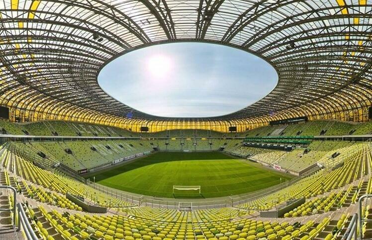 Zdjęcie przedstawia wnętrze stadionu przy pięknej słonecznej pogodzie. Przez kolisty otwór nad boiskiem widać niebieskie niebo. U dołu jest zieleń murawy. W środkowej części zdjęcia znajdują się trybuny dla publiczności, w kolorze żółtym