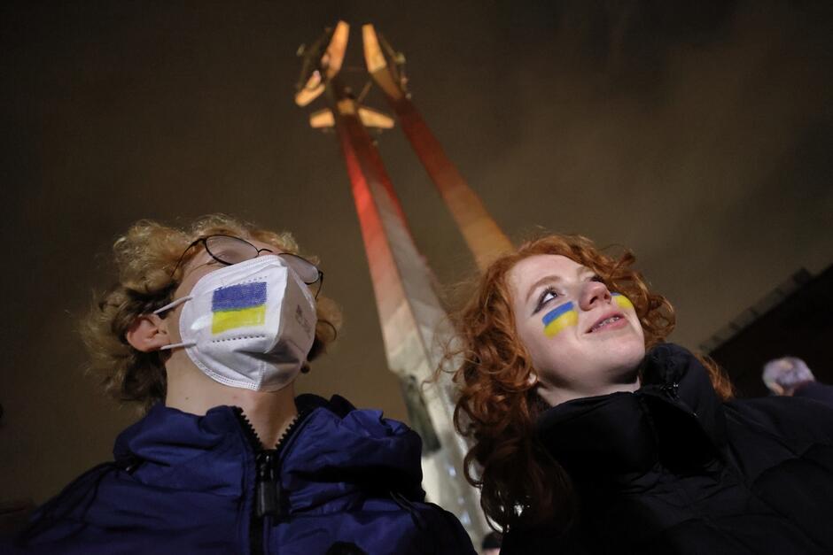 na zdjęciu twarze mężczyzny i kobiety z wymalowanymi na twarzach flagami ukrainy, mężczyzna ma na twarzy białą maseczkę i na niej wymalowaną flagę ukraińską, za głowami widać pomnik poległych stoczniowców w Gdańsku