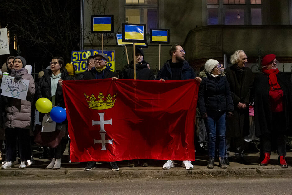 Ciemnawo, kilkanaście osób, kilka trzyma czerwoną flagę z logo Gdańska 