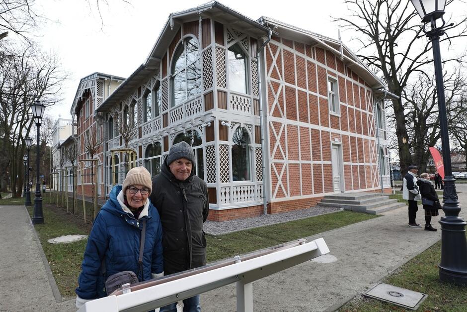 zabytkowy odnowiony budynek, jedna ściana przeszklona, druga z cegły, na pierwszym planie uśmiechnięta para ludzi w średnim wieku