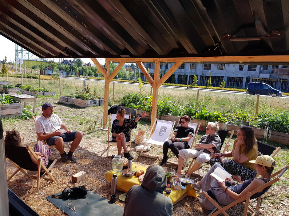 Zdjęcie wykonane na zewnątrz, przedstawia 8 osób siedzących pod wiatą na leżakach wokół stołu, na którym stoją napoje, sałatka w mice, talerzyki, przekąski. W tle znajdują się drewniane rozsadniki z bujnie rosnącymi ziołami, sałatami i warzywami. 
