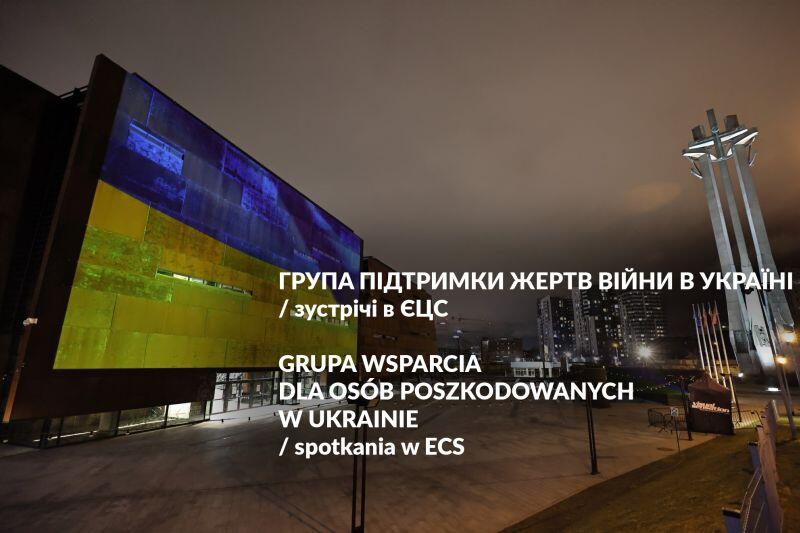 budynek nocą na placu podświetlony na niebiesko-żółto obok trzy krzyże pomnika poległych stoczniowców
