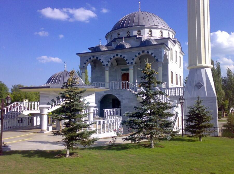 Zdjęcie przedstawia budynek meczetu ze stojącą po prawej świątynną wieżą - minaretem. Cała budowla jest w kolorze białym, znajduje się na posesji pełnej zieleni
