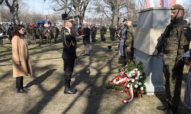 kobieta w płaszczu stoi przed obeliskiem, przed nią salutuje żołnierz, pod pomnikiem wiązanka kwiatów z szarfą biało czerwoną