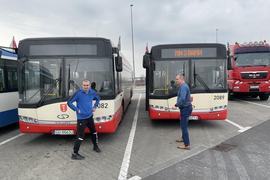 Na zdjęciu są dwa stojące na parkingu autobusy miejskie. Widzimy je od przodu. Przed autobusami stoją dwaj mężczyźni w średnim wieku. Jest to parking przy autostradzie. Na prawo od autobusów widać przód czerwonego tira, który przypadkowo zaparkował obok. Po lewej widać fragment biało-niebieskiego autobusu, który jest w trasie do Lwowa z Gdyni