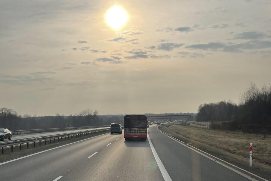 Zdjęcie przedstawia autobusy jadące autostradą, jeden za drugim. Po lewej, na sąsiednim pasie, widać wyprzedzające je auto. Na niebie jest nieco zamglone słońce  