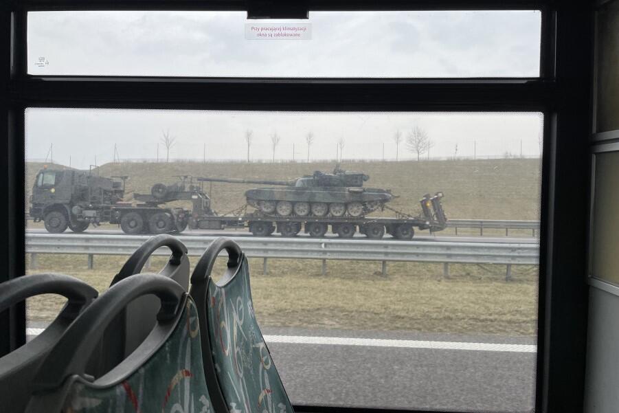 Przez okno autobusu widać ciężarówkę z lawetą, na której transportowany jest czołg. Oba pojazdy pomalowane na ciemnozielony kolor