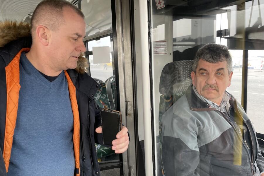 Wnętrze autobusu, widok na kabinę kierowcy. Mężczyzna w średnim wieku stoi po lewej, starszy mężczyzna siedzi po prawej, za kierownicą. Rozmawiają