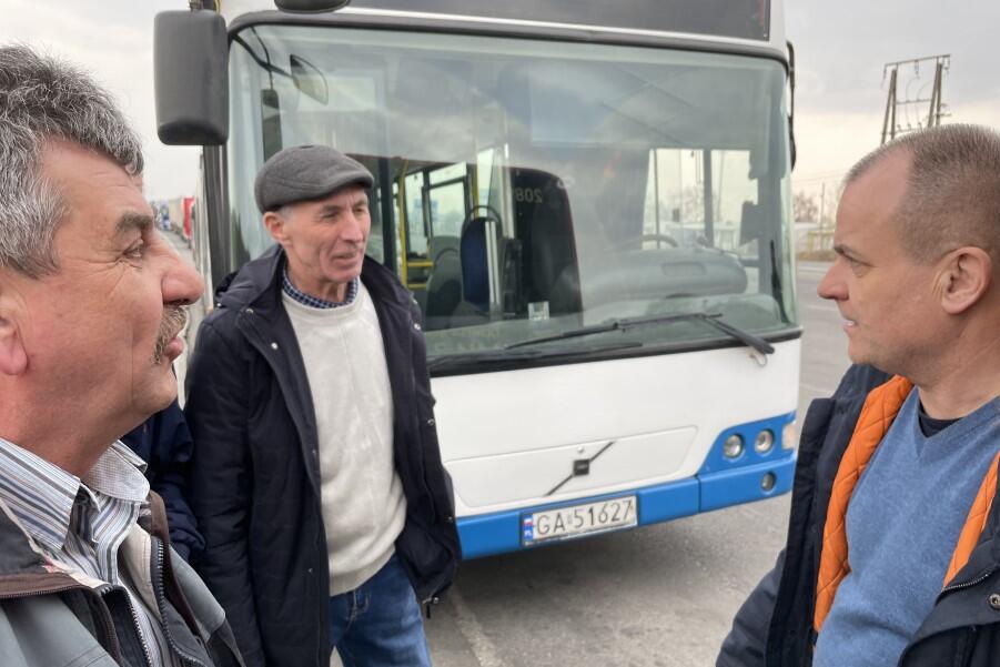 Trzej mężczyźni stoją i rozmawiają przed przodem autobusu. Po prawej jest mężczyzna w średnim wieku, po lewej dwaj starsi mężczyźni - Jarosław i jego kolega w kaszkiecie na głowie