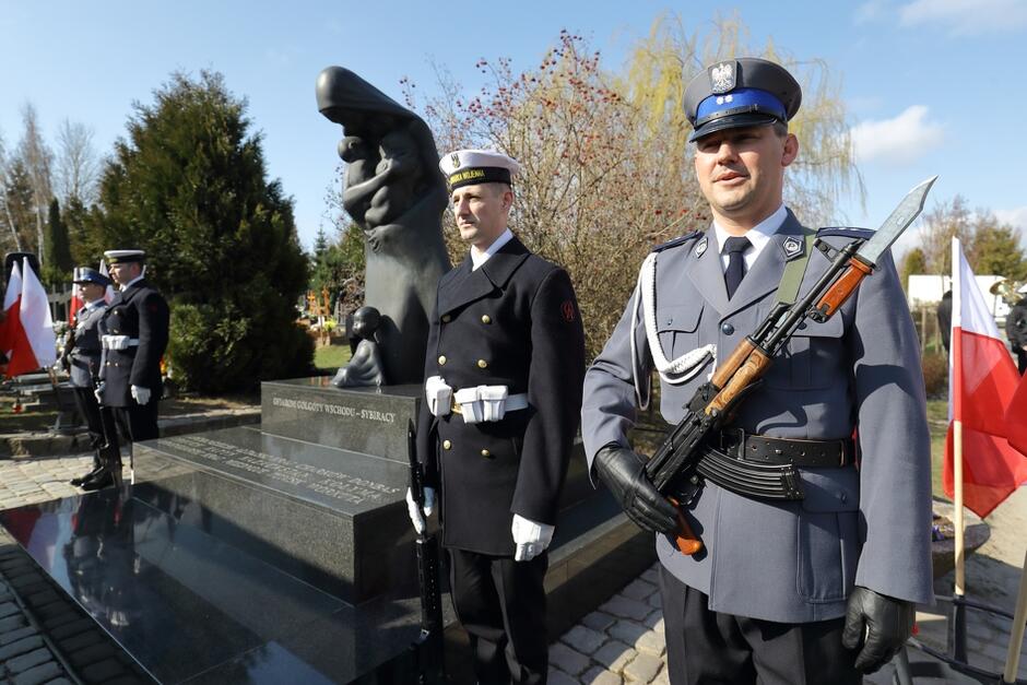 Policjant, marynarze w galowych mundurach z bronią pod pomnikiem. Pomnik to pochylona postać z dzieckiem na ręku. Obok polskie flagi