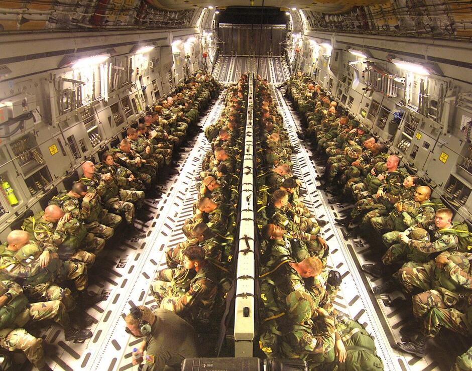 Wnętrze samolotu. Przestronna hala, w której w czterech rzędach siedzą żołnierze w pełnym rynsztunku. Dwa rzędy pod burtami samolotu, dwa rzędy zlokalizowane w środkowej części kadłuba 