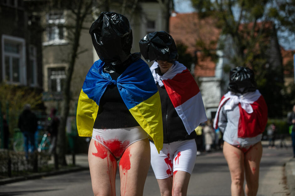 Trzy postacie w workach na głowie z nałożonymi flagami na ramionach (pierwsza ma flagę Ukrainy) w majtkach umazanych sztuczną krwią