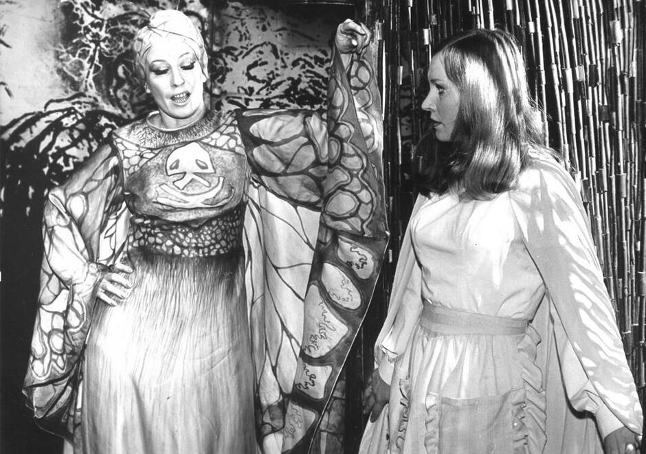 Czarno-białe zdjęcie: dwie kobiety w długich sukniach, po lewej starsza, dłoń na prawym biodrze 