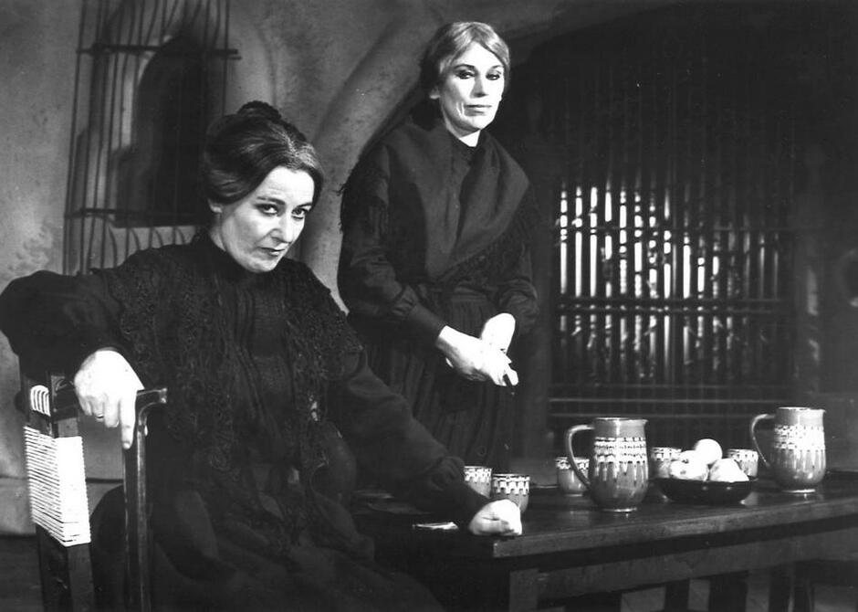 Czarno-białe zdjęcie: dwie kobiety w czarnych szatach, po lewej siedzi i patrzy spod "byka", druga stoi za stołem i patrzy na tę obok