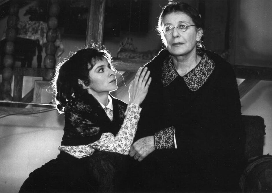 Czarno-białe zdjęcie: dwie kobiety siedzą obok siebie. Po lewej młodsza, dłoń ma opartą i ramię starszej. Starsza w okularach patrzy przed siebie