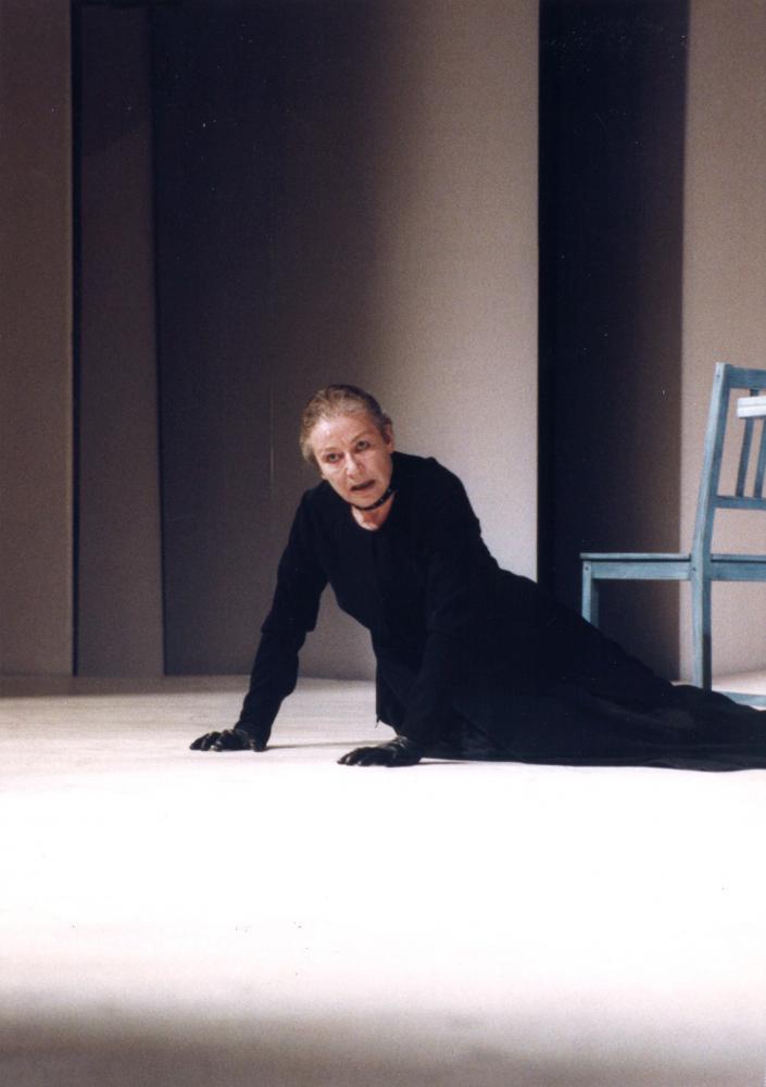 Starsza kobieta w ciemnej sukni siedzi w pozycji półleżącej na podłodze. Za nią ściana i krzesło