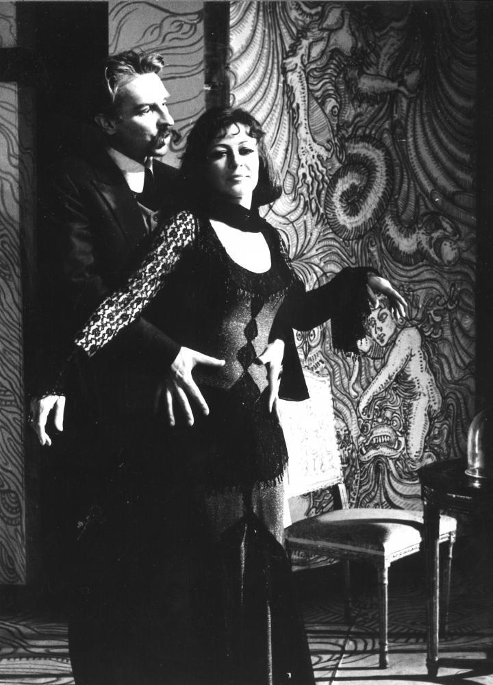 Czarno-białe zdjęcie: mężczyzna i kobieta, on trzyma ręce na jej biodrach. Za nimi krzesło