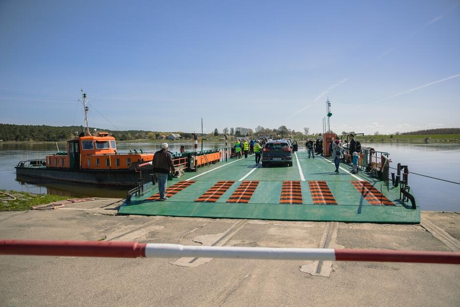 na zdjęciu zielony prom, stoi na nim kilka samochodów osobowych i kilkanaście osób, po lewej widać pomarańczowy mniejszy statek holowniczy, który opiera się o prom