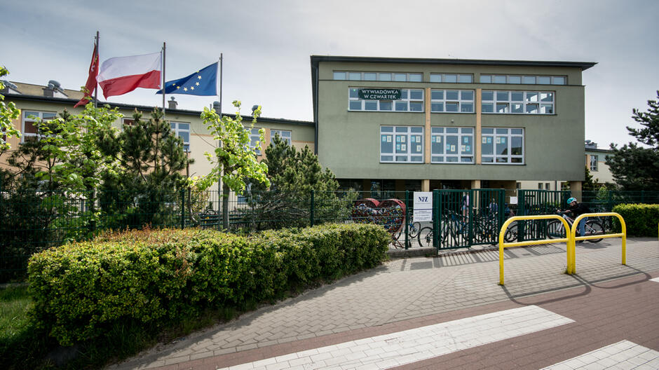 Budynek szkoły, po lewej maszty z flagami Polski i UE, przed budynkiem żywopłot i chodnik
