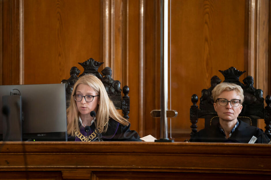 Dwie kobiety w togach sędziowskich siedzą za stołem, po lewej na stole monitor komputera (tyłem do obiektywu aparatu)
