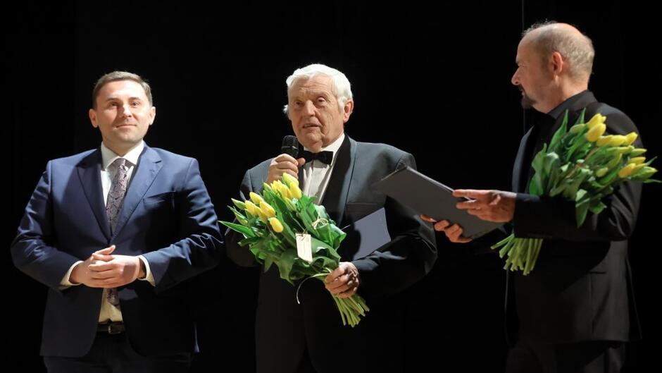 Na scenie stoją trzej mężczyźni w smokingach, dwaj trzymają w rękach bukiety żółtych tulipanów, starszy mężczyzna pośrodku mówi do mikrofonu