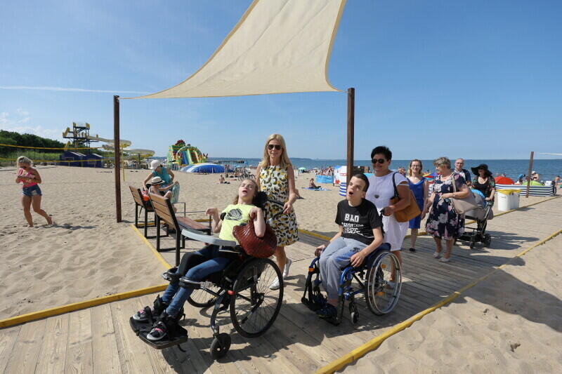 Morski brzeg, woda, piasek, na piasku ułożone chodniki drewnine, po nich jadą nastolatkowie na wózkach inwalidzkich, wiozą je kobiety w średnim wieku.