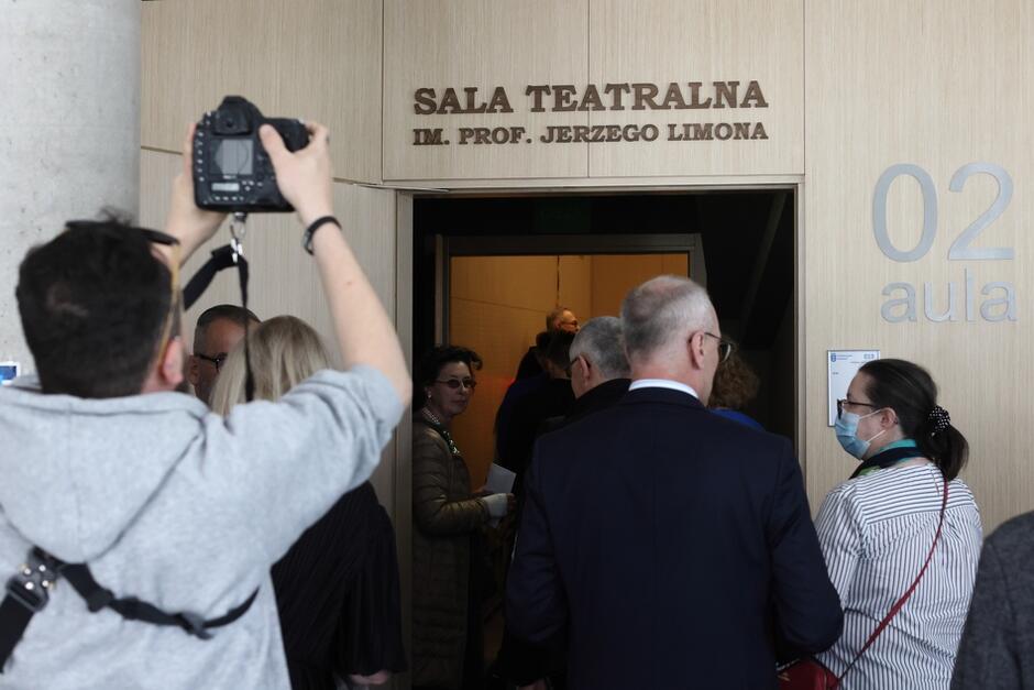 Kilka osób idzie do drzwi wejściowych nad którymi znajduje się napis: Sala Teatralna Imienia Profesora Jerzego Limona