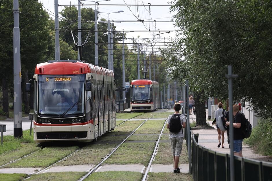 Dwa tramwaje jadące na jednym torze, jeden za drugim. Oba dzieli dystans ponad 100 metrów. Jadą w kierunku widza, nieco w stronę lewego marginesu zdjęcia. Po prawej widać przechodzące osoby. Letnia pora, zielone drzewa