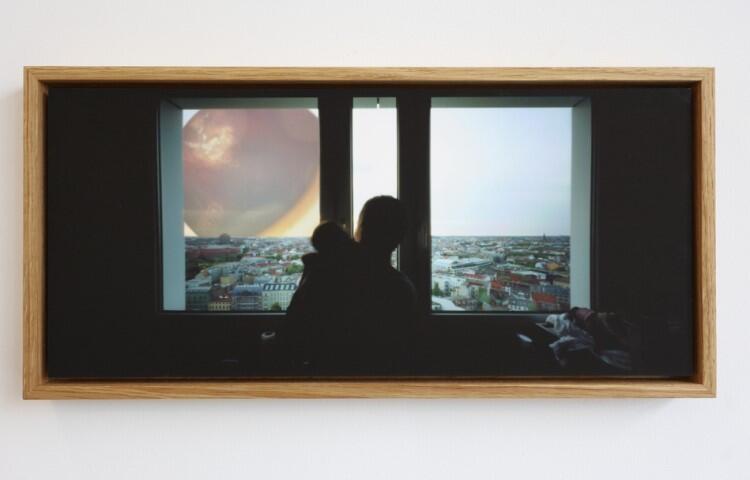 praca artystki: zdjęcie przedstawia kobietę z dzieckiem na ręku patrzących przez okno na miasto