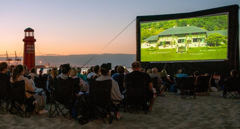 Ekran w trakcie seansów kina letniego na plaży usytuowany jest tyłem do wody, przed nim na krzesłach siedzi publiczność