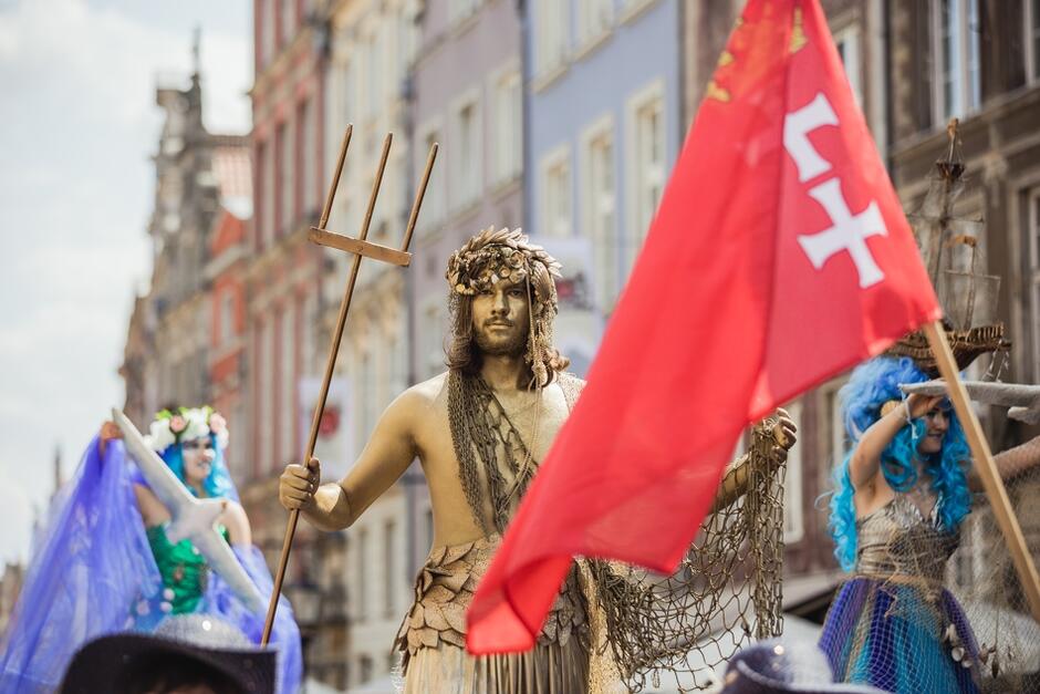 Zdjęcie przedstawia uczestników parady. Jest pomalowana na złoto osoba przebrana w Neptuna z trójzębem i czerwona flaga z herbem Gdańska. Obok Neptuna są jeszcze dwie inne kolorowe - głównie fioletowe - fantazyjne postaci. W tle widzimy kamienice ulicy Długiej