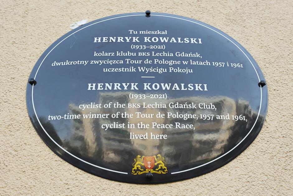 Owalna tablica ciemnego koloru z białymi napisami, informującymi kim był Henryk Kowalski - po polsku i angielsku. U dołu tablicy znajduje się herb Miasta Gdańska z lwami 