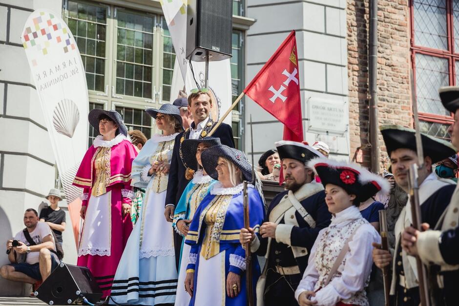 Przedproże Dworu Artusa, stoją na nim mężczyźni i kobiety w strojach z epoki, trzymają flagę Gdańska 