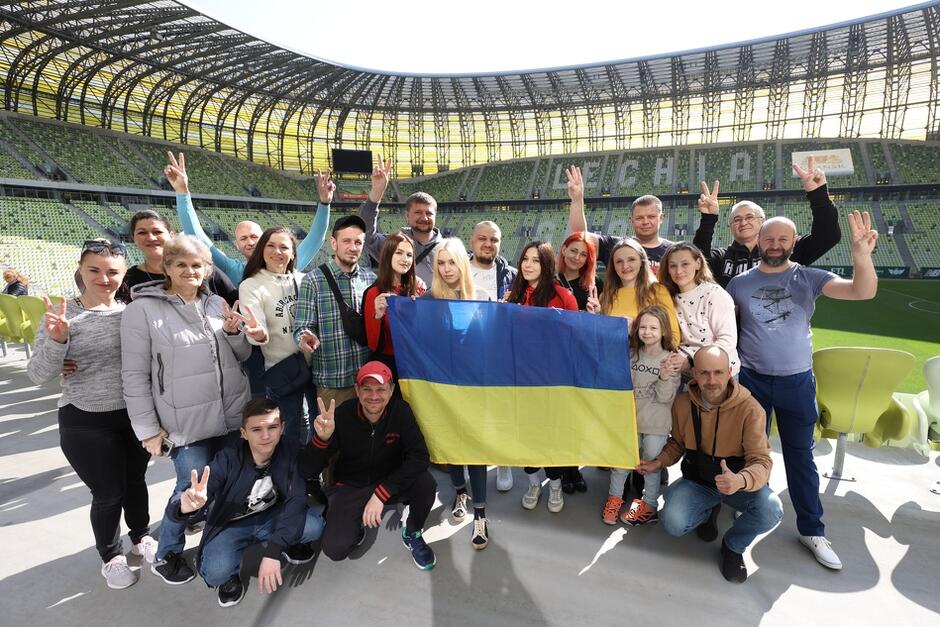 Stadion, stoi kilkadziesiąt osób, kobiet i mężczyzn w różnym wieku, dzieci. Część się uśmiecha, macha, kilka osób trzyma flagę ukraińską 