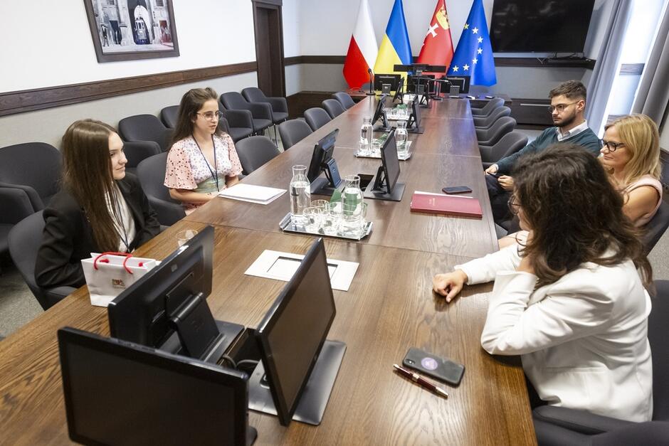 Duży stół, na pierwszym planie laptopy, w tle flagi Polski, Ukrainy, Unii Europejskiej, z lewej strony siedzą dwie dziewczyny, z lewej dwie kobiety w średnim wieku i młody mężczyzna