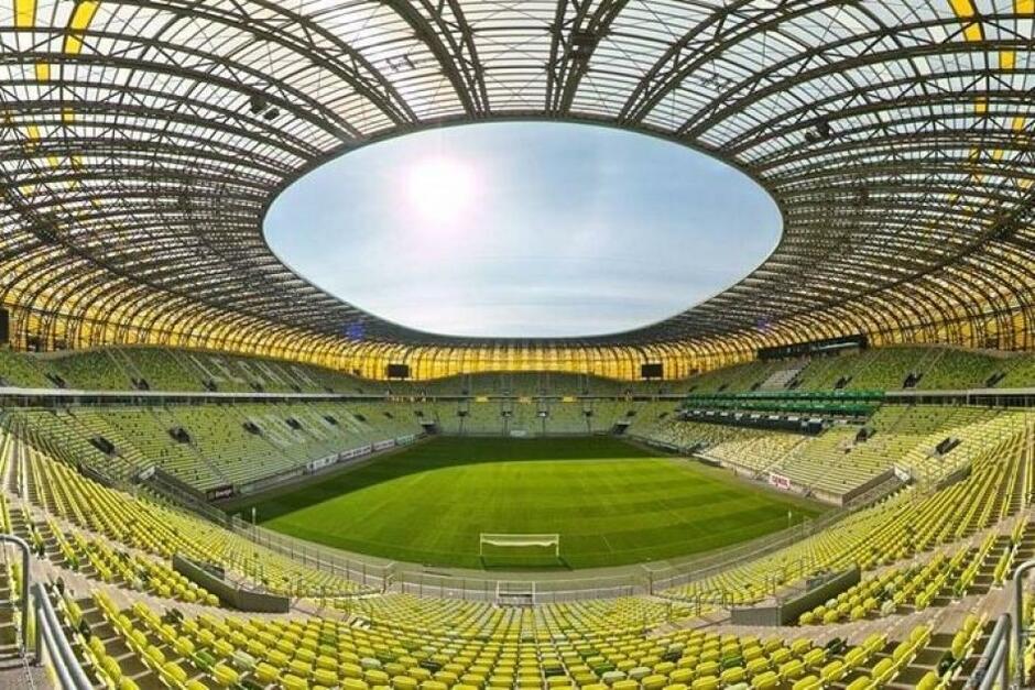 Wnętrze stadionu pokazane tzw. żabim okiem, przy dziennym świetle. Dominują odcienie zieleni i kolor żółty, przechodzący w bursztynowy. Widać murawę, trybuny, koronę stadionu i pogodne niebo ponad obiektem 